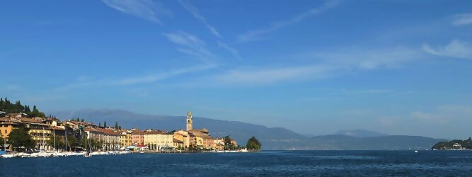 Trip Voucher beliebte Urlaubsziele am Gardasee -  Mit einer Fläche von 370 km² ist der Gardasee der größte See Italiens. Es liegt am Fuße der Alpen und erstreckt sich über drei Staaten: Lombardei, Venetien und Trentino. Die maximale Tiefe des Sees beträgt 346 m, er hat eine längliche Form und sein nördliches Ende ist sehr schmal. Dort ist der See von den Bergen der Gruppo di Baldo umgeben. Du trittst aus deinem gemütlichen Hotelzimmer und es begrüßt dich die warme italienische Sonne. Du blickst auf den atemberaubenden Gardasee, der in zahlreichen Blautönen schimmert - von tiefem Dunkelblau bis zu funkelndem Türkis. Majestätische Berge umgeben dich, während die Brise sanft deine Haut streichelt und der Duft von blühenden Zitronenbäumen deine Nase kitzelt. Du schlenderst die malerischen, engen Gassen entlang, vorbei an farbenfrohen, blumengeschmückten Häusern. Vereinzelt unterbricht das fröhliche Lachen der Einheimischen die friedvolle Stille. Du fühlst dich wie in einem Traum, der nicht enden will. Jeder Schritt führt dich zu neuen Entdeckungen und Abenteuern. Du probierst die köstliche italienische Küche mit ihren frischen Zutaten und verführerischen Aromen. Die Sonne geht langsam unter und taucht den Himmel in ein leuchtendes Orange-rot - ein spektakulärer Anblick.